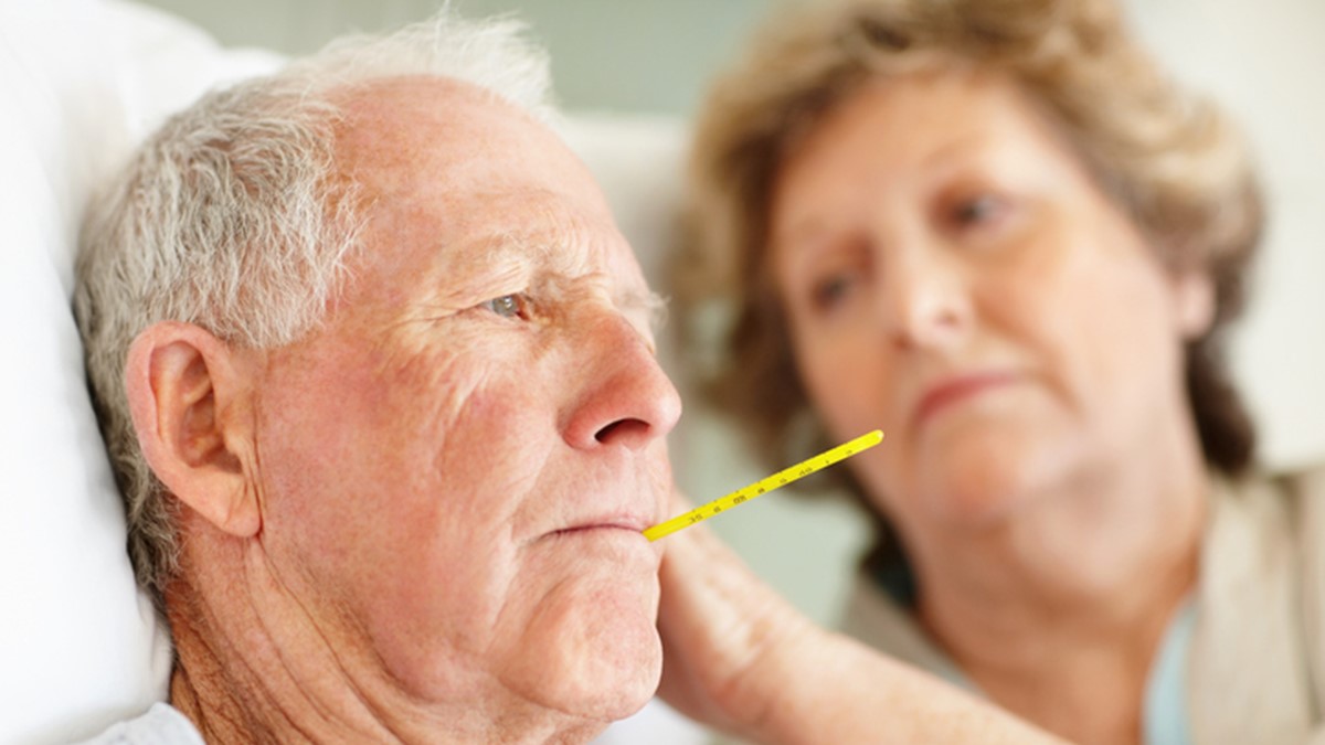 Older male sick in bed with thermometer in mouth; older female watches.