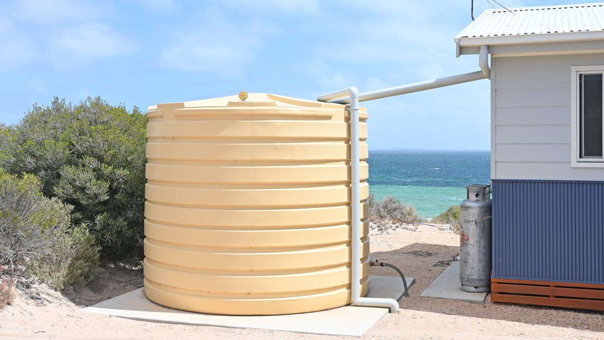 Large, plastic water storage tank (cistern) on a concrete pad. A plastic pipe connects the cistern to a house. Another plastic pipe goes from the cistern into the ground. The cistern and house are overlooking the ocean.