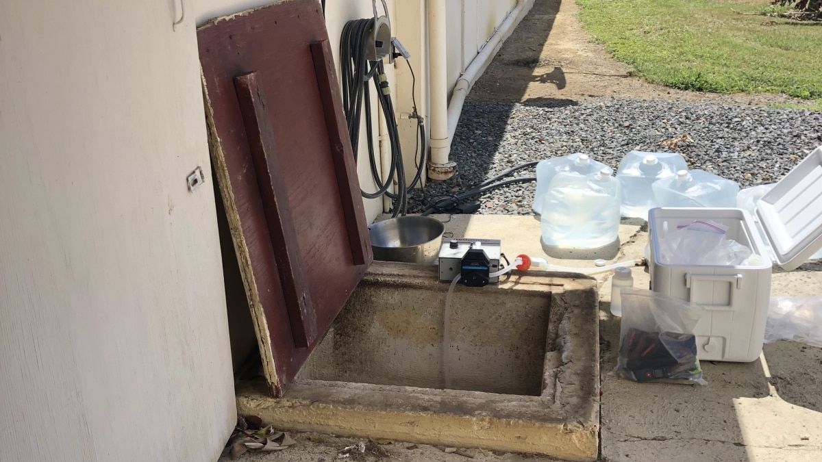 An underground cistern outside next to a building. The wooden lid of the cistern is up, showing a square concrete opening. A cooler, bags of water, and a testing instrument are around the cistern opening.